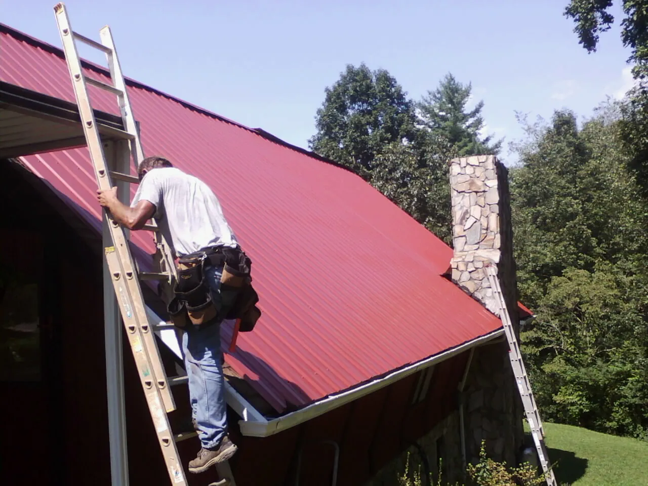 Roofer on a ladder beside a red metal roof and stone chimney — Soffit & Fascia Repair by Roofing Brothers in Mount Joy